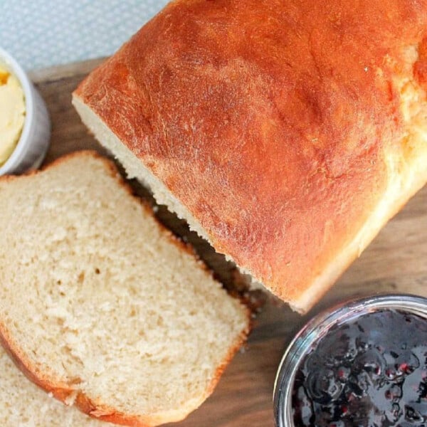 Loaf of bread with slices cut, showcasing the art of baking bread at home, sits beside butter and a jar of jam on a wooden board.