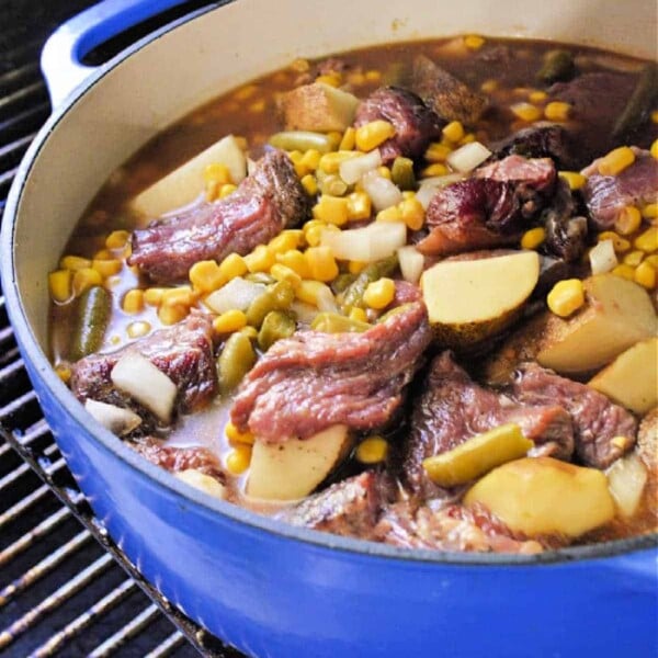 Blue pot with a smoky stew of beef stew meat, potatoes, corn, and diced vegetables simmering on the grill.