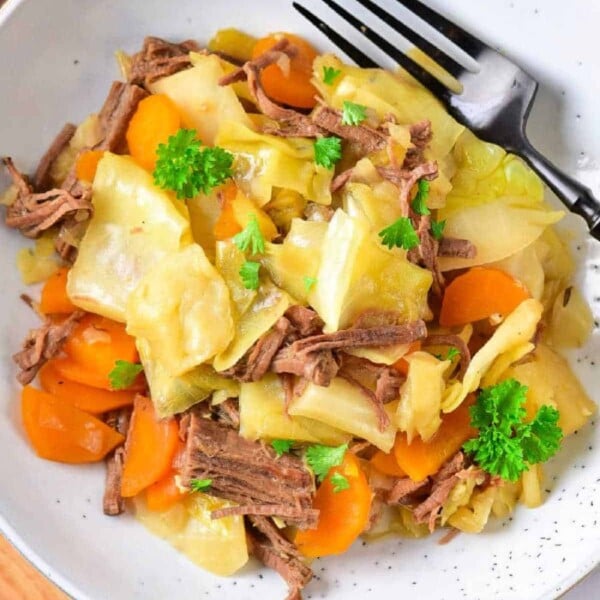 A plate of beef with tender cabbage stew, carrots, and parsley graces a light-colored plate alongside a fork.